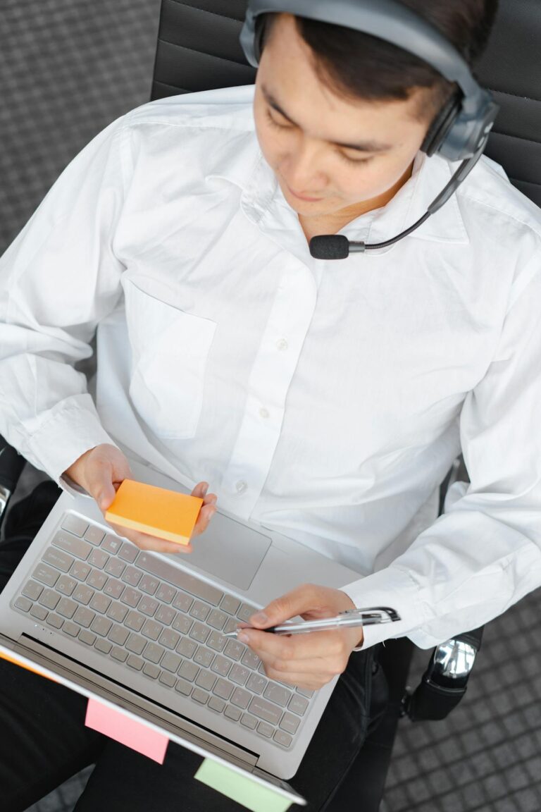 Man in white shirt multitasking with headset and laptop for call center work.